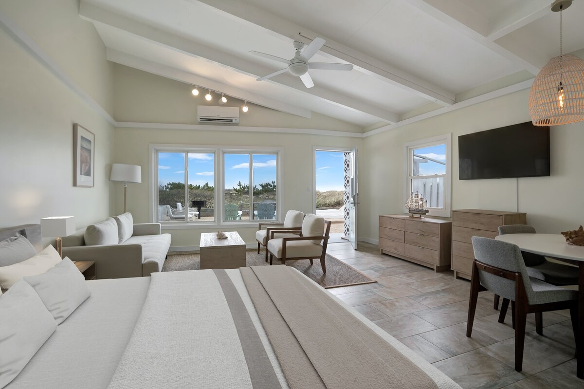 A bright studio unit features high ceilings and large windows that provide views of the outdoor landscape. A spacious bed is positioned in the foreground, with a cozy seating area and a dining table in the living space. Light-colored furnishings complement the neutral decor.