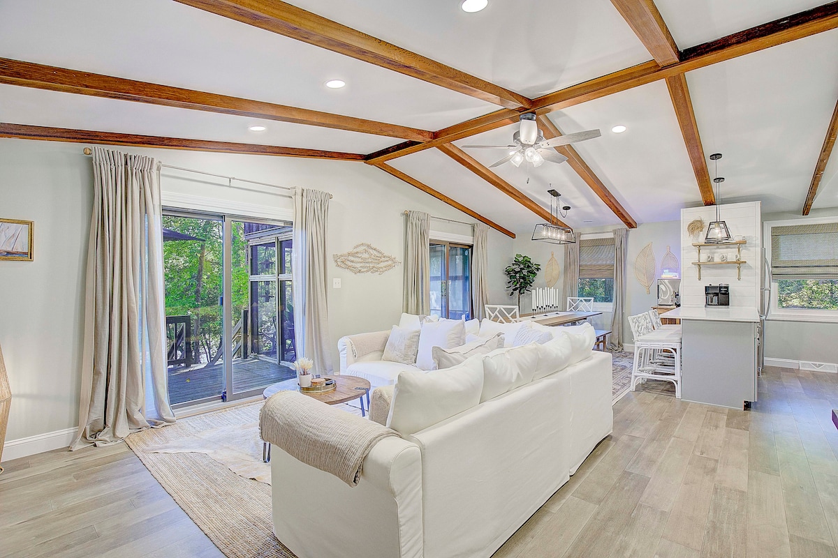 A bright living area is highlighted by wooden beam ceilings and light-colored furnishings. Large sliding glass doors offer views of the outdoors, while natural light fills the space. A comfortable seating arrangement provides a welcoming environment, complemented by soft textiles and a neutral color palette.