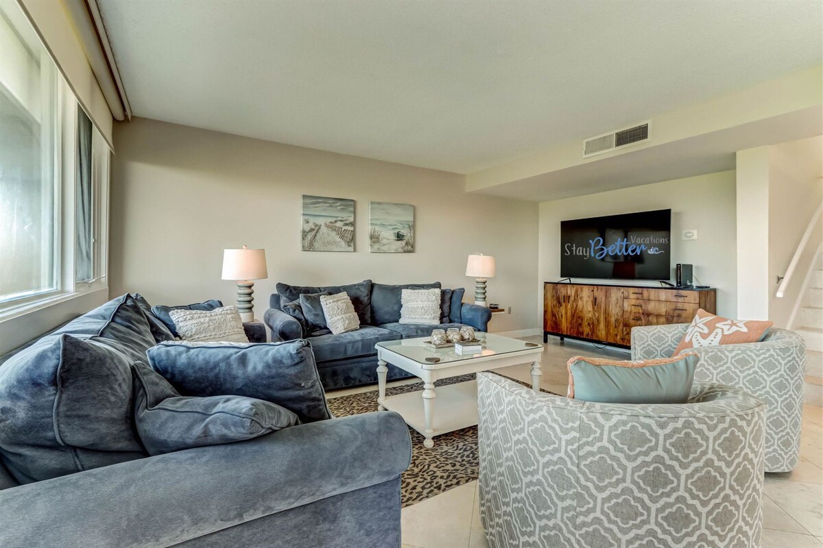 A spacious living room is furnished with multiple soft sofas and chairs, offering ample seating for guests. A large TV is mounted on the wall, while a decorative cabinet adds style. Natural light enters through the windows, enhancing the relaxed atmosphere.