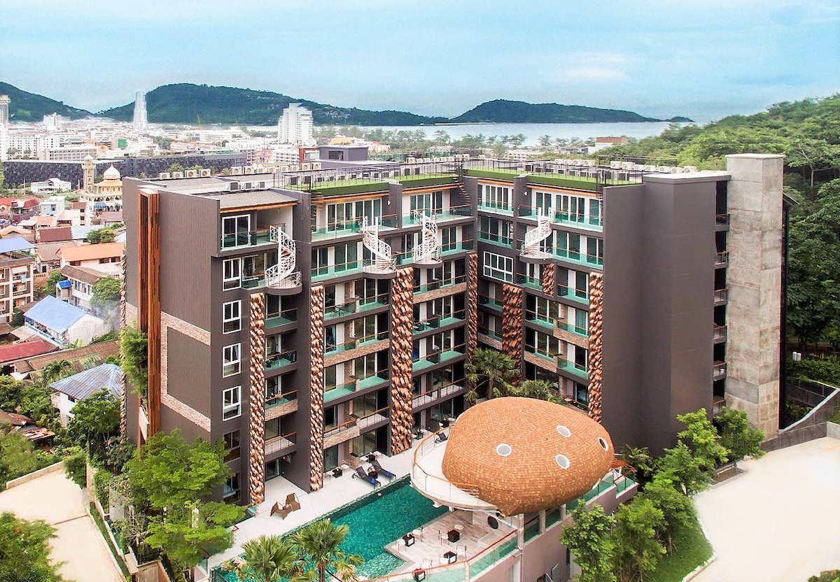 An aerial view of the Emerald Terrace Resort showcases a modern, multi-story building surrounded by lush greenery. A communal swimming pool is visible at the forefront, complemented by a unique mushroom-shaped structure nearby. The backdrop includes distant hills and the coastal landscape beyond.