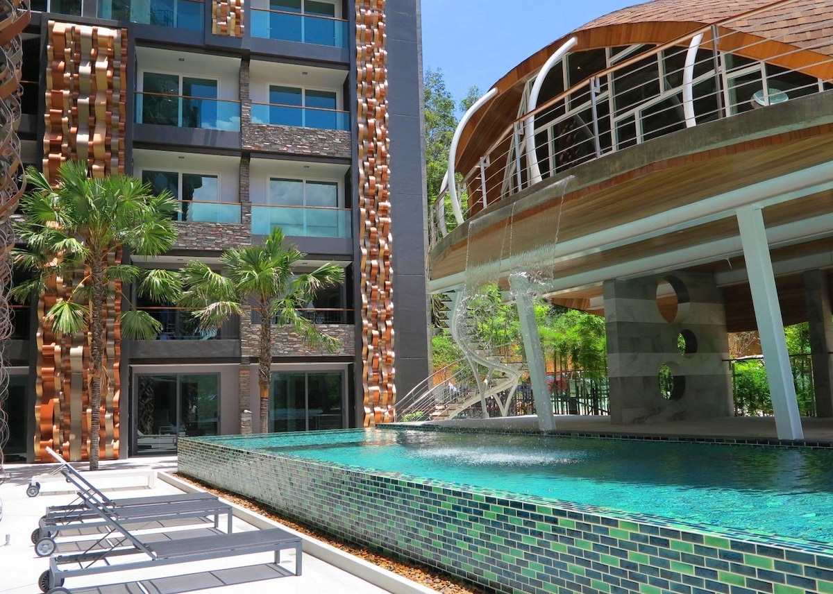 A serene communal area showcases an infinity pool with a mosaic tile design and cascading water features. Surrounding palm trees add a touch of greenery, while modern architecture provides a relaxing backdrop. Lounge chairs are positioned near the pool for guest enjoyment.