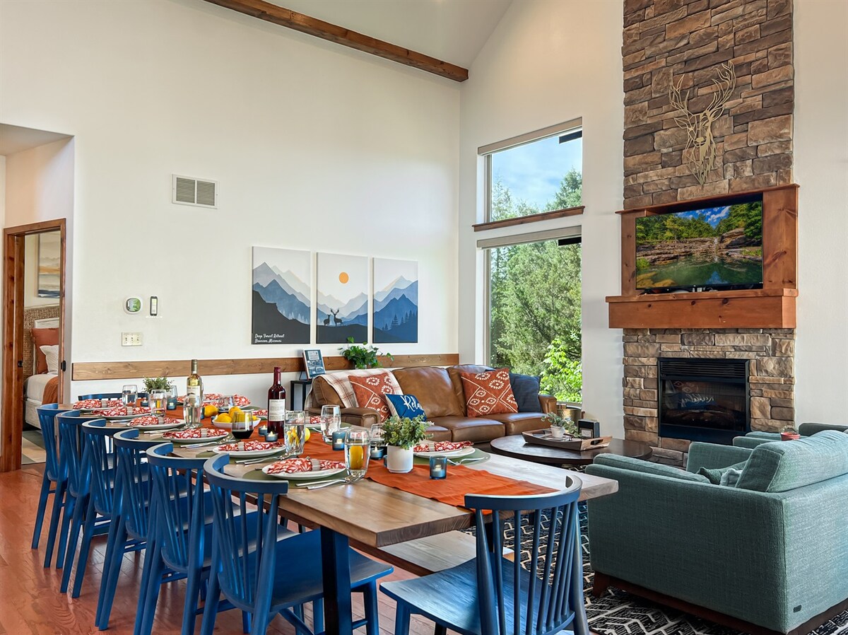 A spacious dining area features a large wooden table set for a meal, accommodating multiple guests. The inviting living room includes a comfortable sofa with decorative pillows, a stone fireplace, and large windows that provide views of the surrounding forest.