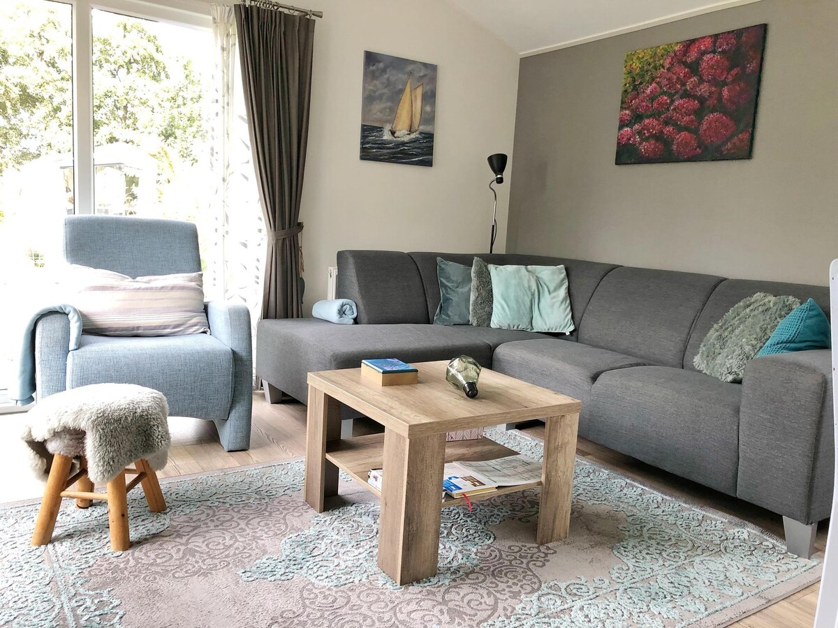 The modern living area features a comfortable gray sectional sofa and an accent chair next to a large window. A wooden coffee table sits centrally, adorned with a decorative stone and magazines. Soft light filters through the curtains, creating a cozy atmosphere.