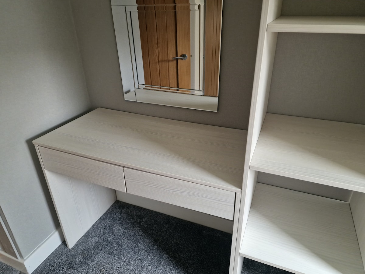 A light-colored vanity desk is positioned against a wall, featuring two drawers for storage. Above the desk, a rectangular mirror is mounted. A nearby shelf unit displays open space, providing additional storage options, and the floor is covered with grey carpeting.