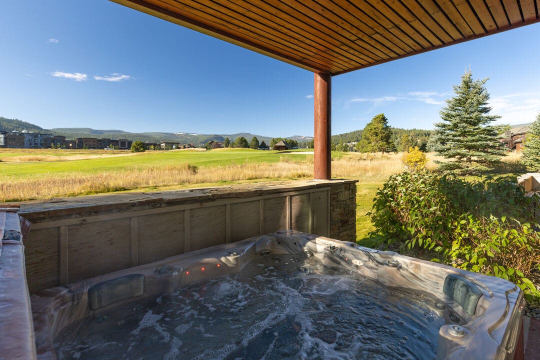 A private hot tub is positioned under a wooden overhang, offering views of a grassy expanse that leads to the Big Sky Golf Course. The clear blue sky and distant mountains create a serene backdrop, inviting relaxation in this picturesque outdoor space.