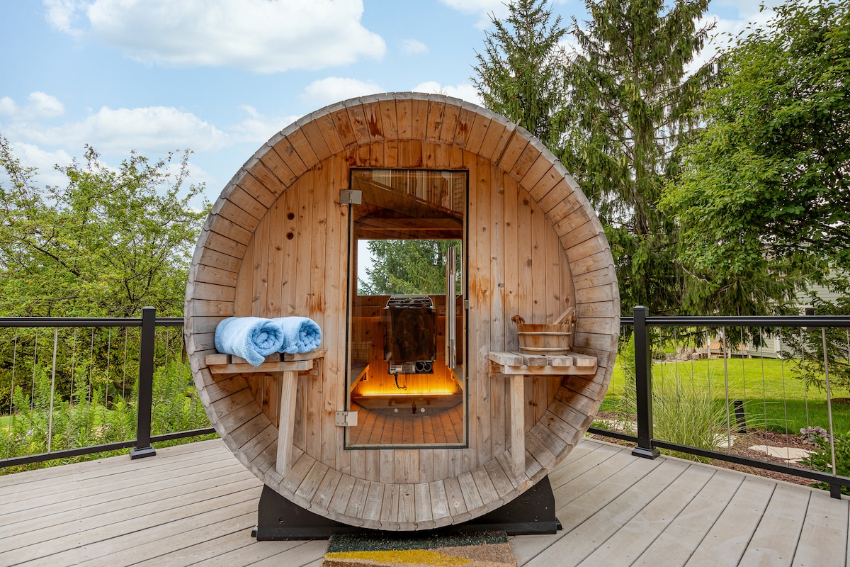 An outdoor cedar barrel sauna is situated on a spacious deck, featuring a round wooden design with an entrance door. Shelves are built into the interior walls, holding towels and essential items. Lush greenery surrounds the sauna, enhancing the natural setting.