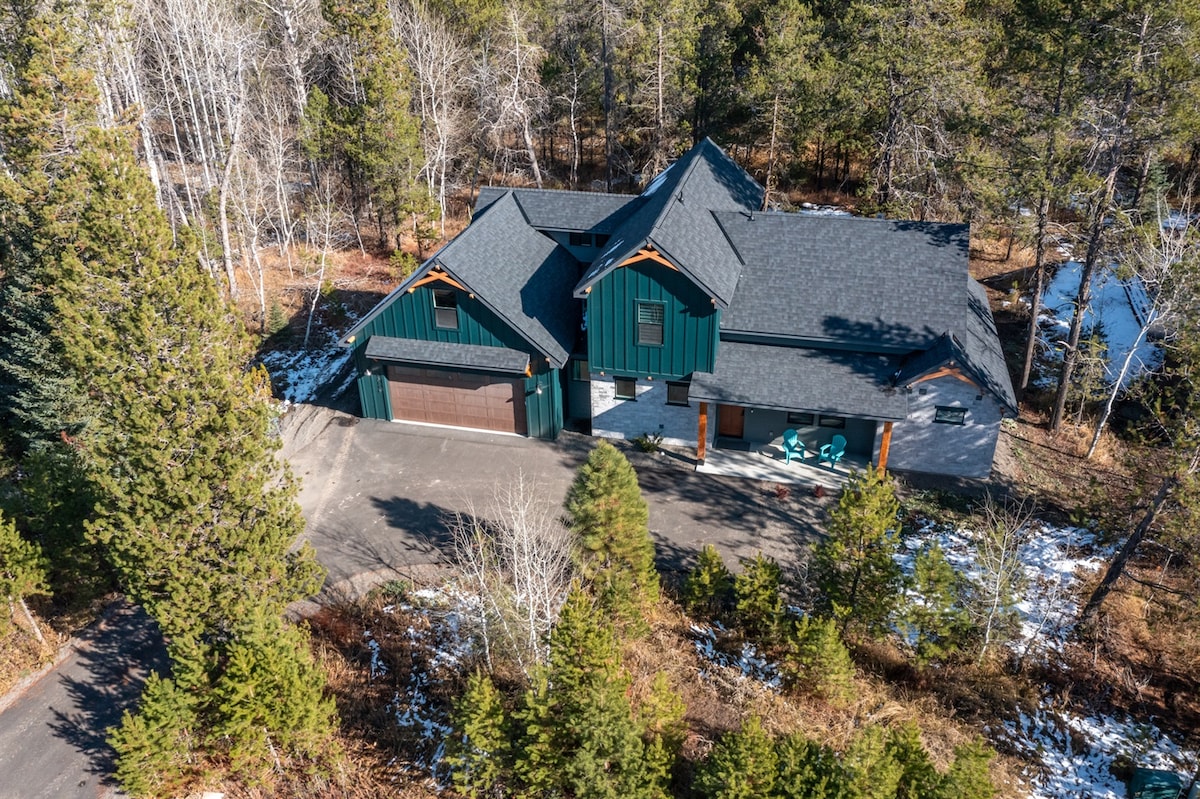 An aerial view of a spacious cabin surrounded by trees and greenery. The structure features a dark green exterior with contrasting wooden accents and a large driveway leading to the garage. Snow patches are visible on the ground, indicating a mountain environment.