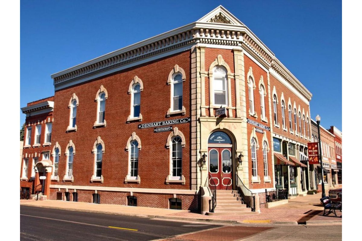 An elegant brick building showcases architectural details from the 1870s, featuring arched windows and ornate trim. A welcoming entrance is framed by decorative wall signage, while shopfronts line the lower level, reflecting the charm of a historic small-town setting.