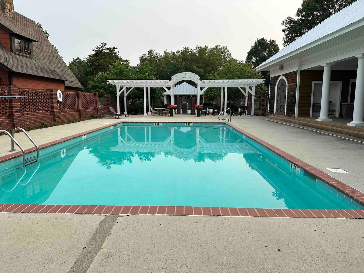 A clear swimming pool is shown with turquoise water, surrounded by a spacious deck. Nearby, a pavilion with seating can be seen, accompanied by shaded areas adorned with tables and chairs, offering a relaxing outdoor space. Lush greenery frames the perimeter.