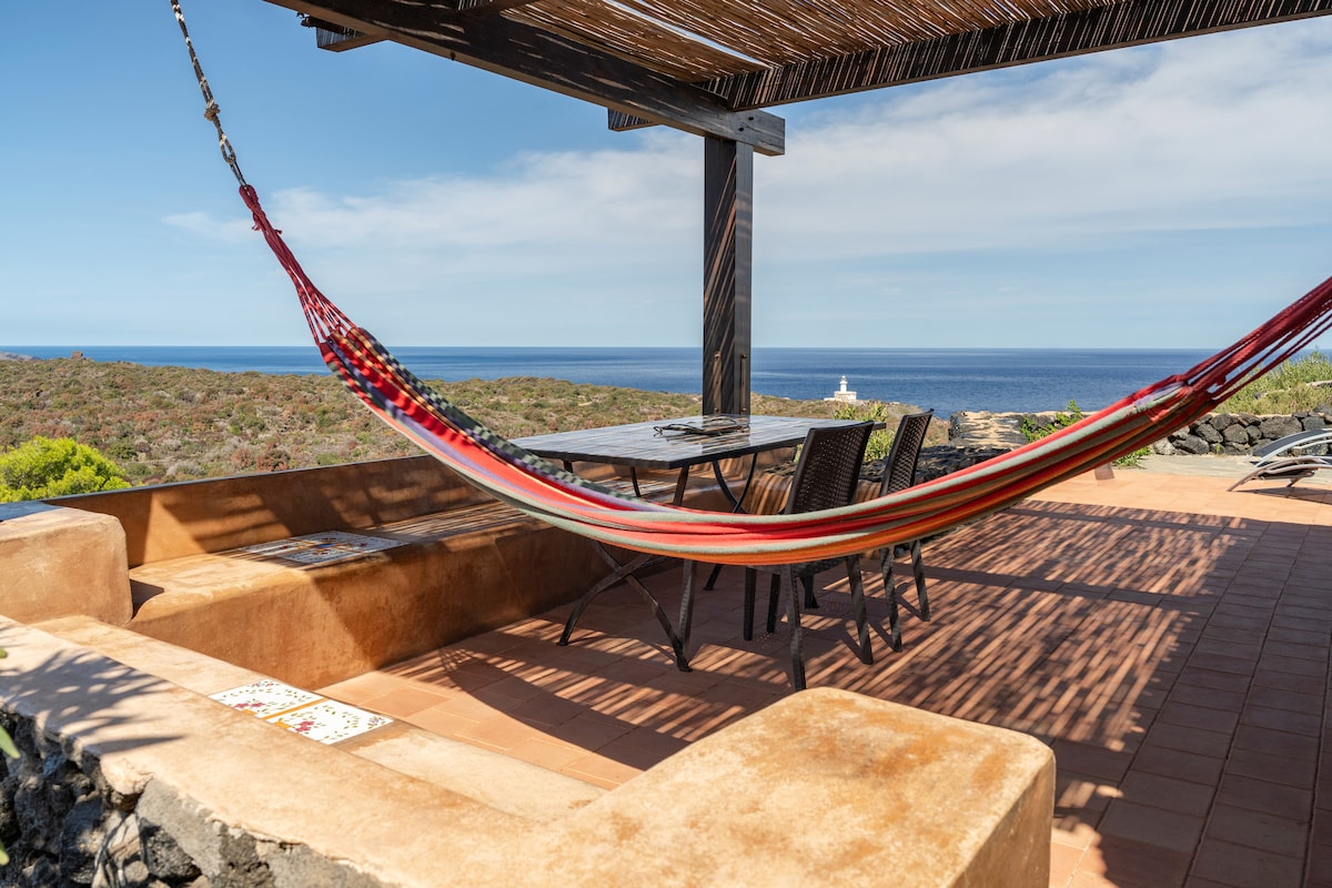 An outdoor terrace is equipped with a dining table and seating, inviting relaxation. A colorful hammock is suspended between wooden posts, overlooking the expansive sea view. Natural light filters through the canopy above, creating a serene environment.