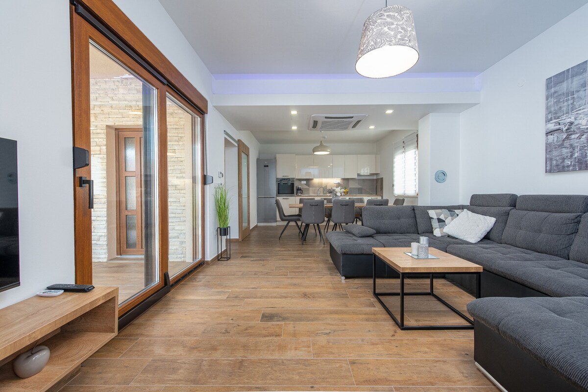 A modern living area features a spacious sectional sofa and a coffee table, situated on a wooden floor. Large glass sliding doors provide access to an outdoor space, while a dining table and kitchen are visible in the background. Soft lighting enhances the inviting atmosphere.