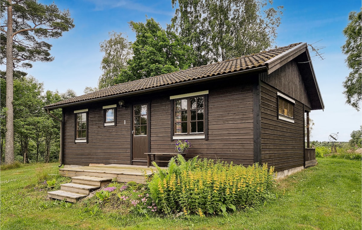 A charming wooden cottage is nestled among lush greenery, featuring a sloped roof and a welcoming front porch. Large windows allow natural light to fill the interior, while a wooden bench sits on the porch, creating a cozy outdoor space.