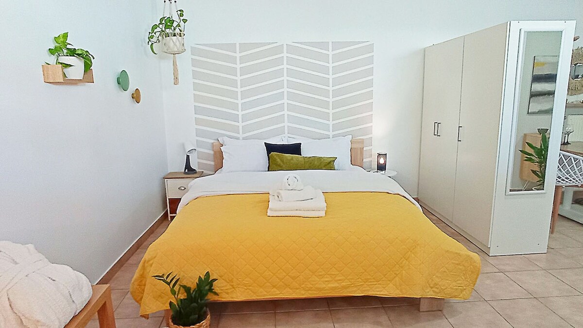 A spacious sleeping area features a yellow quilted bedspread draped over a large bed positioned against a decorative wall with a chevron pattern. Potted plants are displayed on nearby shelves, and a white storage cabinet stands to the side, enhancing the calming atmosphere.