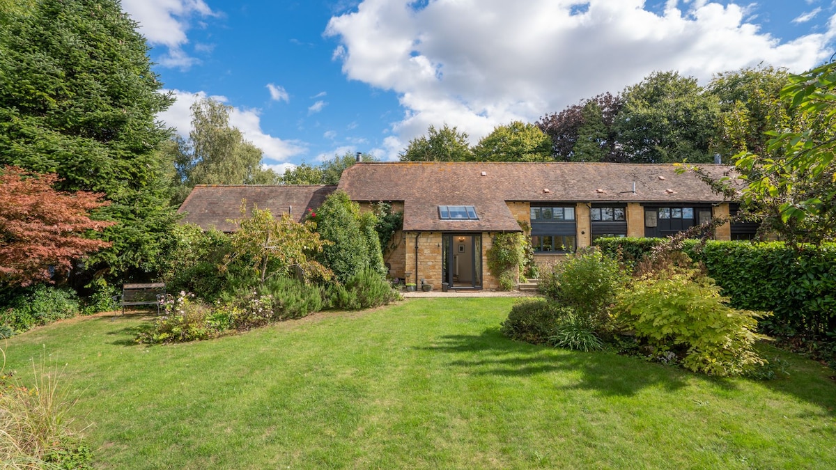 A spacious garden surrounds the converted Cotswold stone barn, featuring well-maintained lawns and a variety of greenery. Natural light casts a warm glow on the exterior, with the barn’s charming architecture highlighted against the backdrop of trees and a partially cloudy sky.