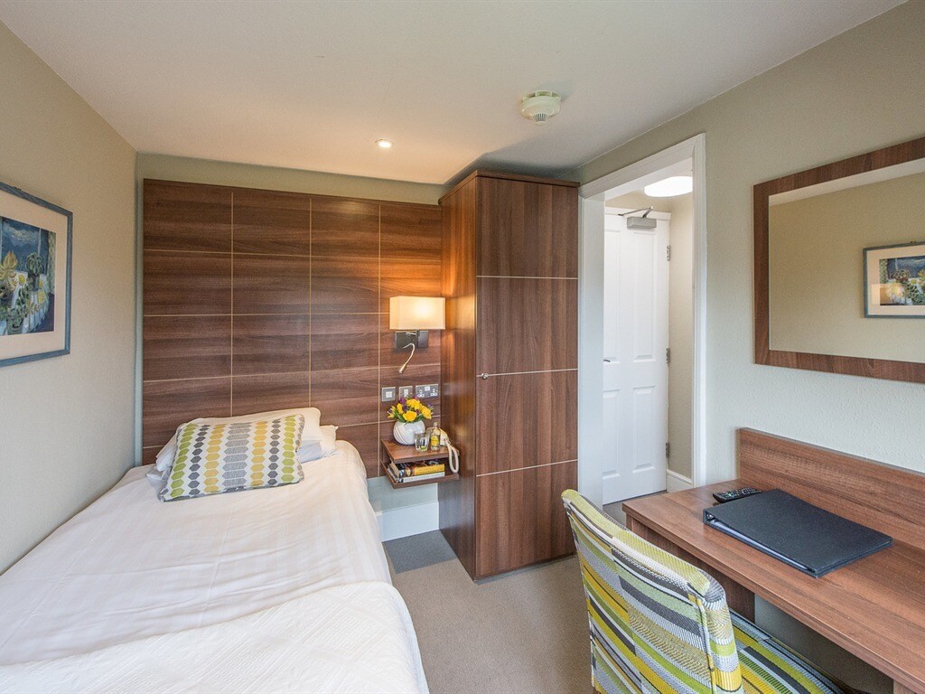 A small ensuite room features a single bed with a patterned throw and a bedside table with fresh flowers. A compact workspace with a chair and desk is present. Built-in storage cabinets are against one wall, and a mirror reflects the room's natural light.