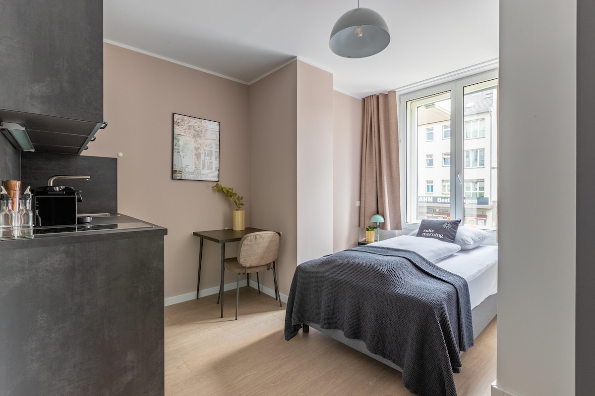 A bright room is featured with large windows allowing natural light to enter. A single bed is dressed with dark bedding, and a compact dining area is situated nearby. A kitchenette with modern finishes and subtle decor elements complete the inviting living space.