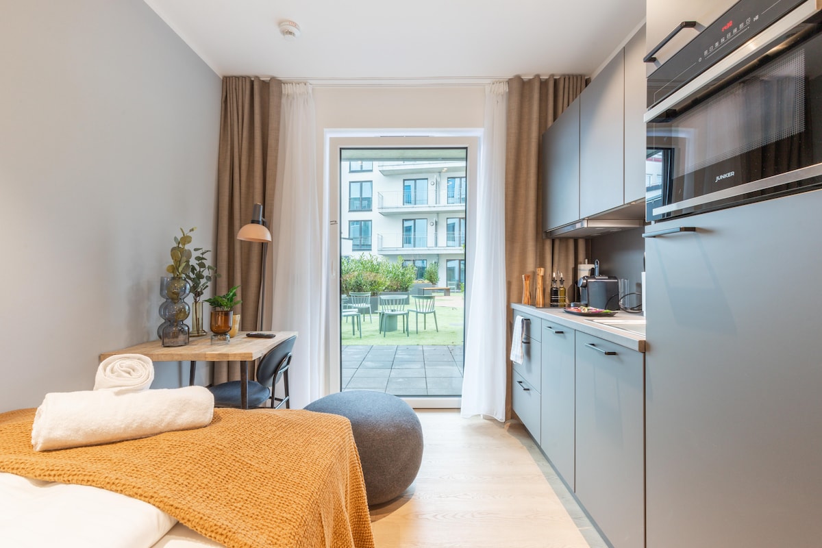 A compact living space is highlighted by a cozy bed with a warm blanket. A small dining table is arranged next to a large window, allowing natural light to illuminate the room. The kitchenette features modern appliances, while a serene outdoor view is visible through the patio door.