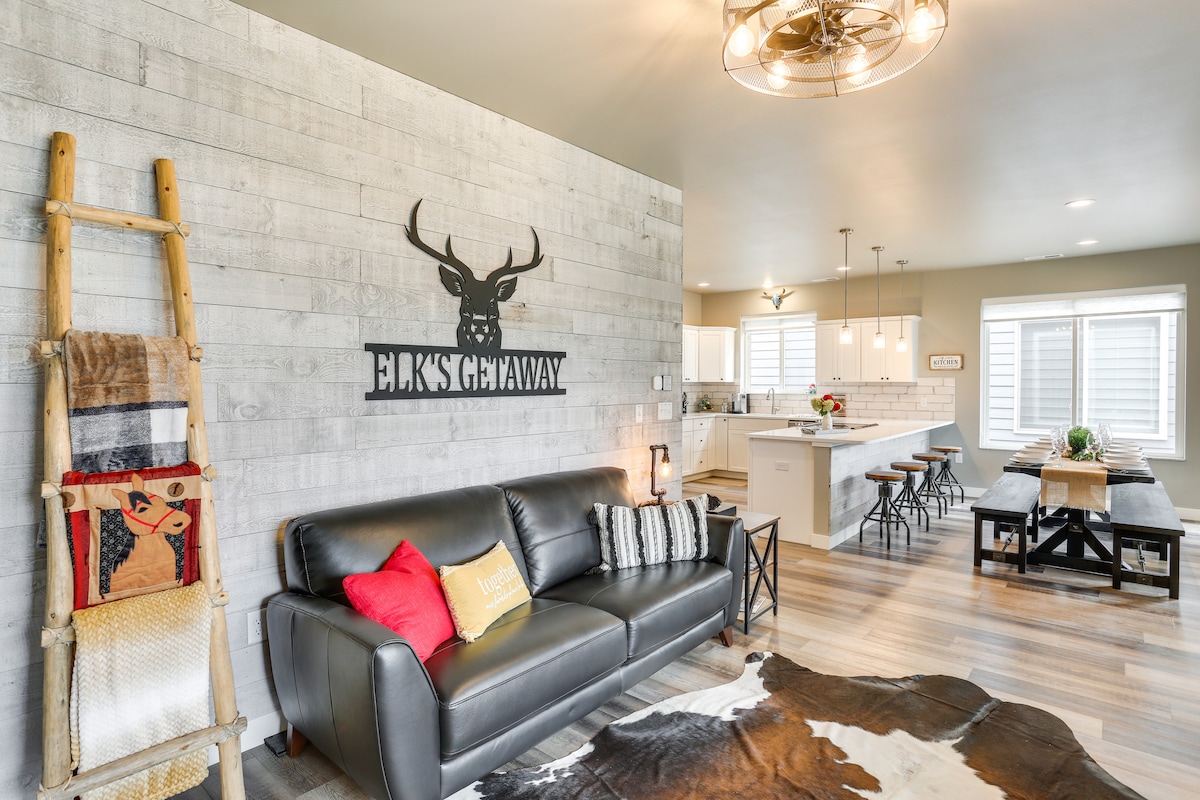 A modern living space features a leather sofa adorned with decorative pillows. A rustic accent wall displays the phrase 'Elk's Getaway.' A large dining table and kitchen area are visible in the background, complemented by pendant lighting and large windows allowing natural light.