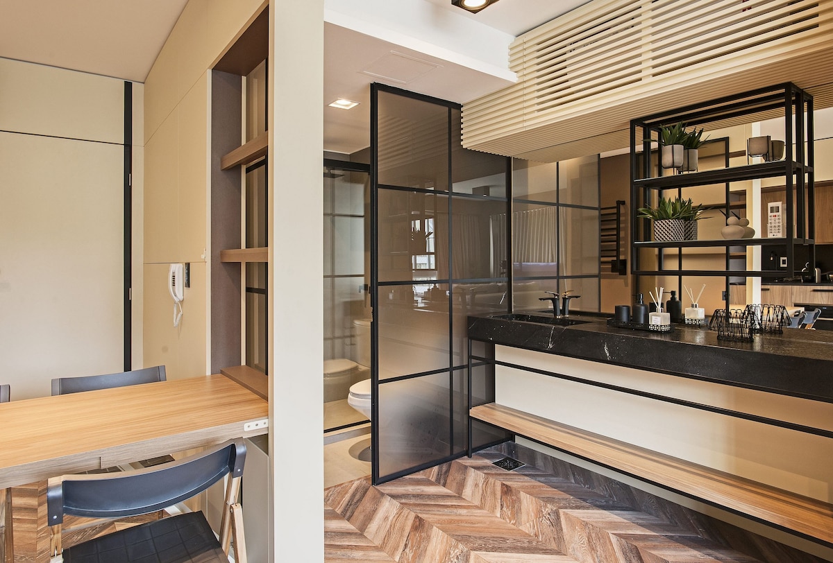 A modern kitchen space is visible, featuring a black countertop and open shelving. Natural wood dining table and chairs are positioned nearby. Glass partitions separate the kitchen from additional areas, allowing for light to fill the space. Stylish decor and plants add a clean, contemporary touch.