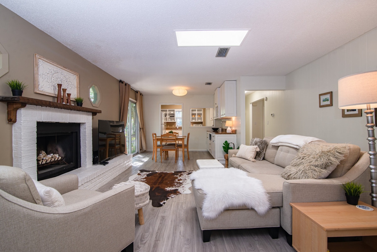 A spacious living area features a light-colored sofa and armchair, arranged around a neutral rug. A brick fireplace adds a cozy touch while a dining area is visible in the background. Natural light filters in through large windows, enhancing the calm atmosphere.