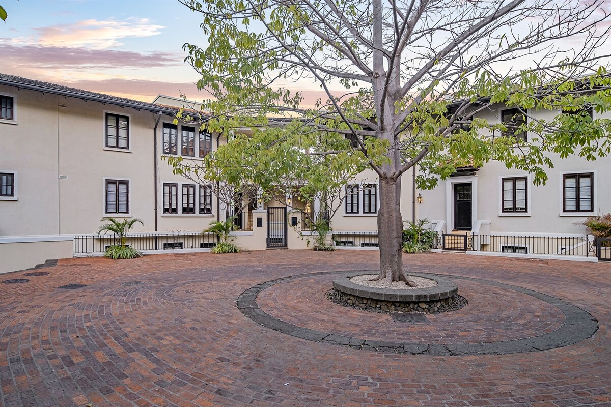 A well-maintained courtyard features a central tree, surrounded by a circular brick pathway. The building is accented by multiple large windows, enhancing the inviting atmosphere. Lush plants line the perimeter, contributing to the serene environment within the community.
