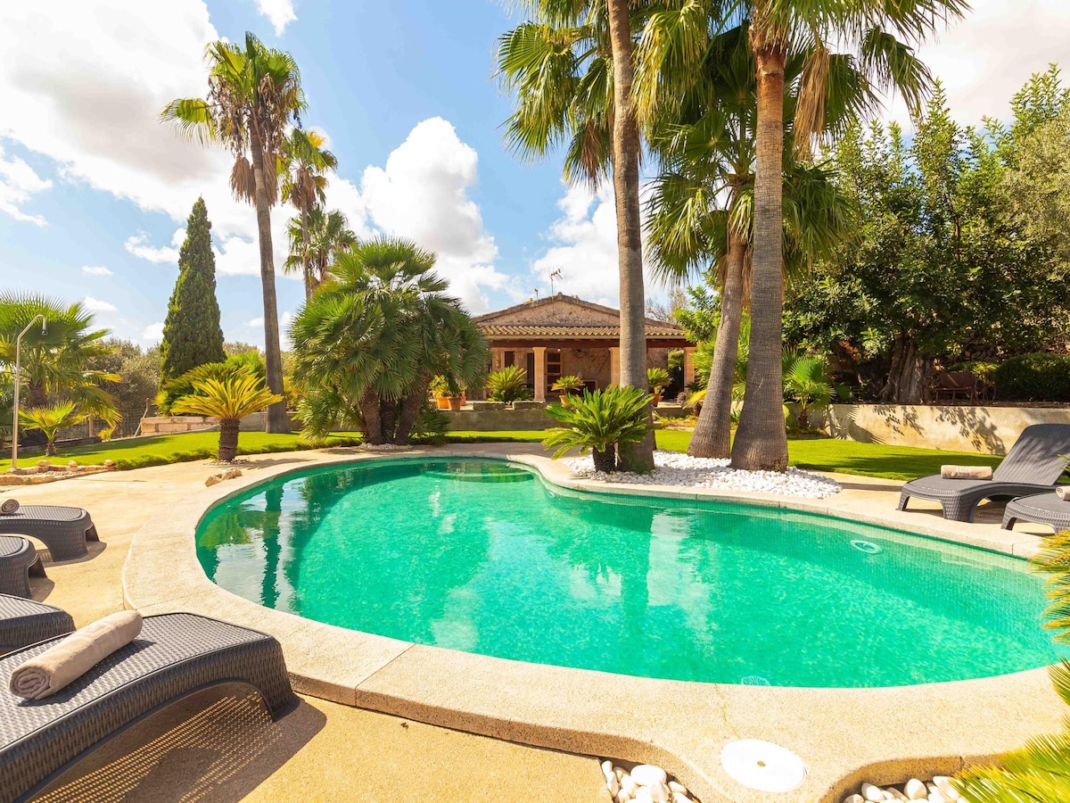 A private pool is surrounded by palm trees and sun loungers. The crystal-clear water reflects the blue sky and fluffy clouds above. A rustic villa is seen in the background, framed by lush greenery and landscaped gardens.
