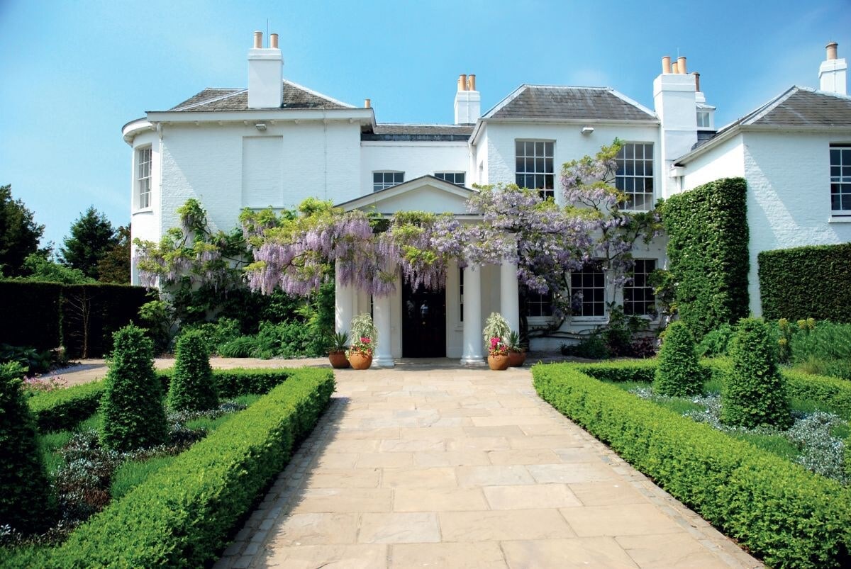 A white, two-story house is framed by lush greenery and flowers. A stone pathway leads to the entrance, which is adorned with ornamental planters. Climbing plants enhance the facade, creating an inviting appearance amid well-maintained hedges and garden areas.