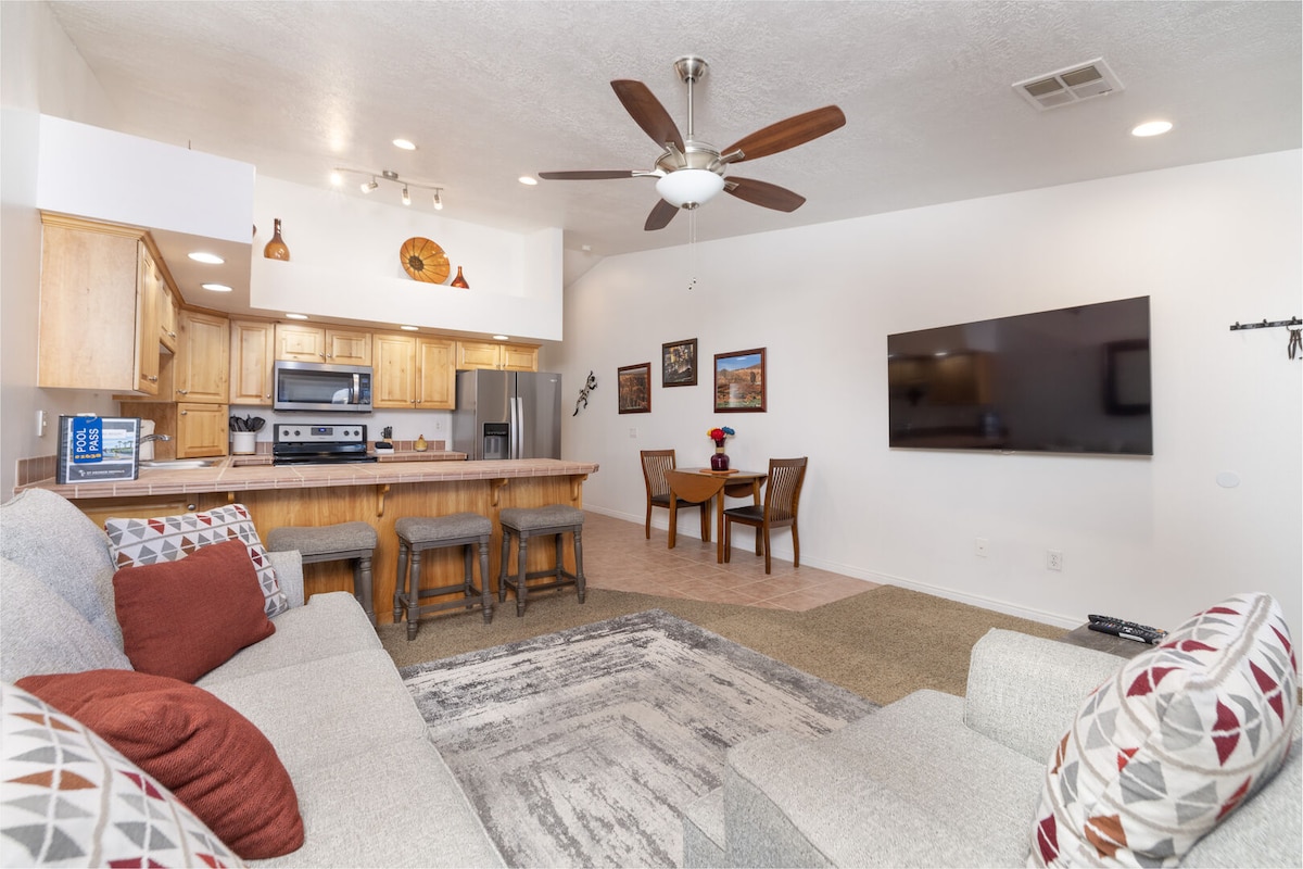 The open living area features a cozy sectional sofa adorned with decorative pillows, a wooden dining table with chairs, and a modern kitchen with stainless steel appliances. A large wall-mounted television is visible, and natural light fills the space through multiple windows.