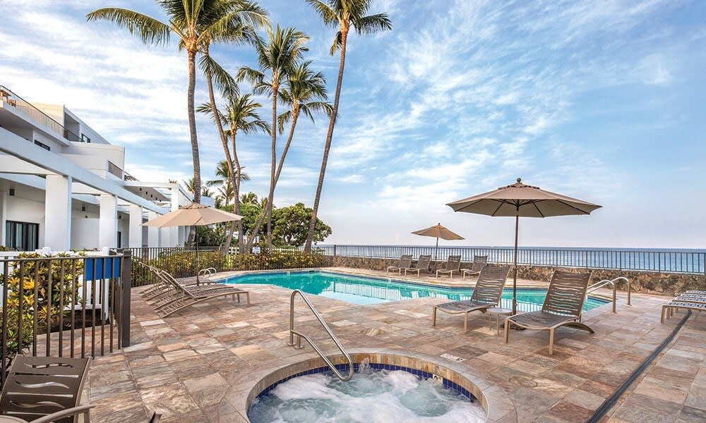 An outdoor pool area is surrounded by palm trees, with lounge chairs arranged around the shimmering water. An inviting hot tub is positioned nearby, offering relaxation. Umbrellas provide shade for guests while ocean views are visible in the distance.