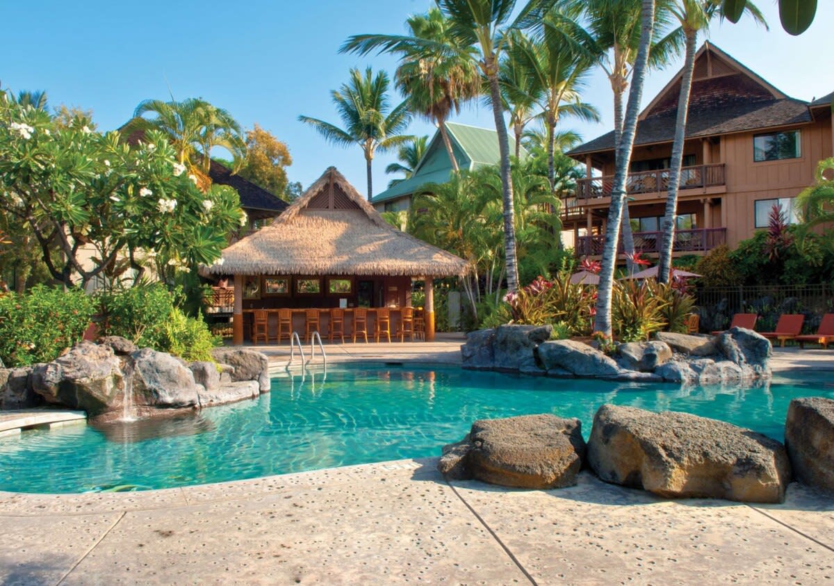 A serene outdoor pool is framed by lush palm trees and tropical foliage. Natural rock formations surround the water, creating a harmonious space. A thatched roof structure can be seen in the background, alongside a building featuring balconies and colorful landscaping.
