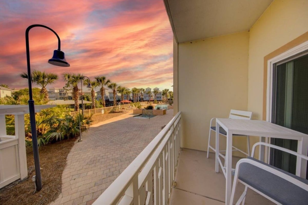 A private balcony is showcased with white outdoor furniture facing a scenic view of a landscaped area. A vibrant sunset illuminates the sky in hues of orange and pink, while palm trees and a community pool are visible in the background.