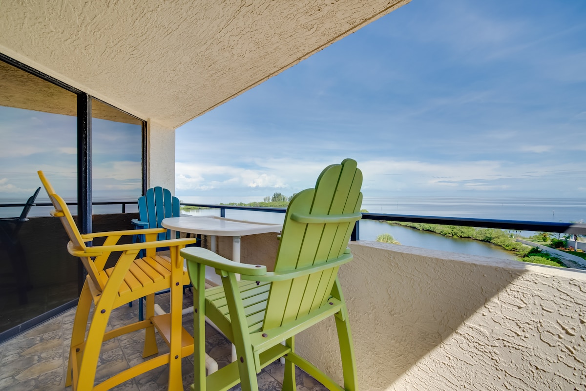 A private balcony is showcased with colorful high-back chairs in yellow and green, accompanied by a small round table. The railing offers an unobstructed view of the surrounding landscape and the water, reflecting the calmness of the scenery.