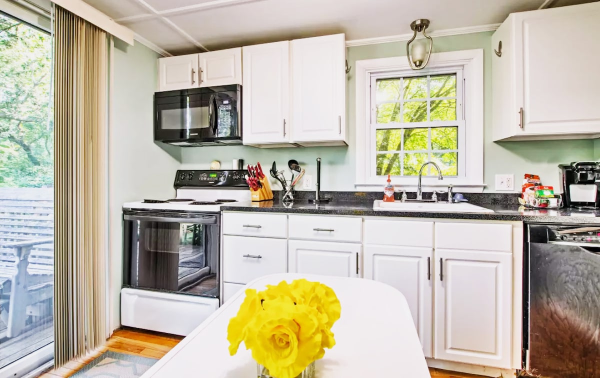 A bright kitchen features white cabinetry and a dark countertop, showcasing modern appliances including a stove, microwave, and refrigerator. A small table is centered with a vibrant bouquet of yellow flowers, adding a touch of color. Natural light filters through a nearby window, illuminating the space.