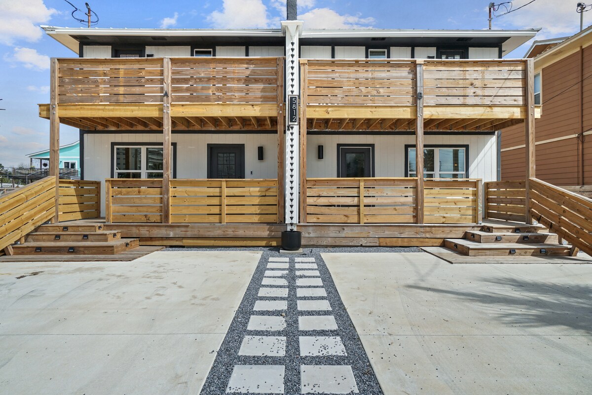 The exterior showcases a two-story townhome with a welcoming wooden deck featuring railings on both levels. A stone pathway leads to the entrance, flanked by wooden steps and a gravel border, providing a charming approach to the home.