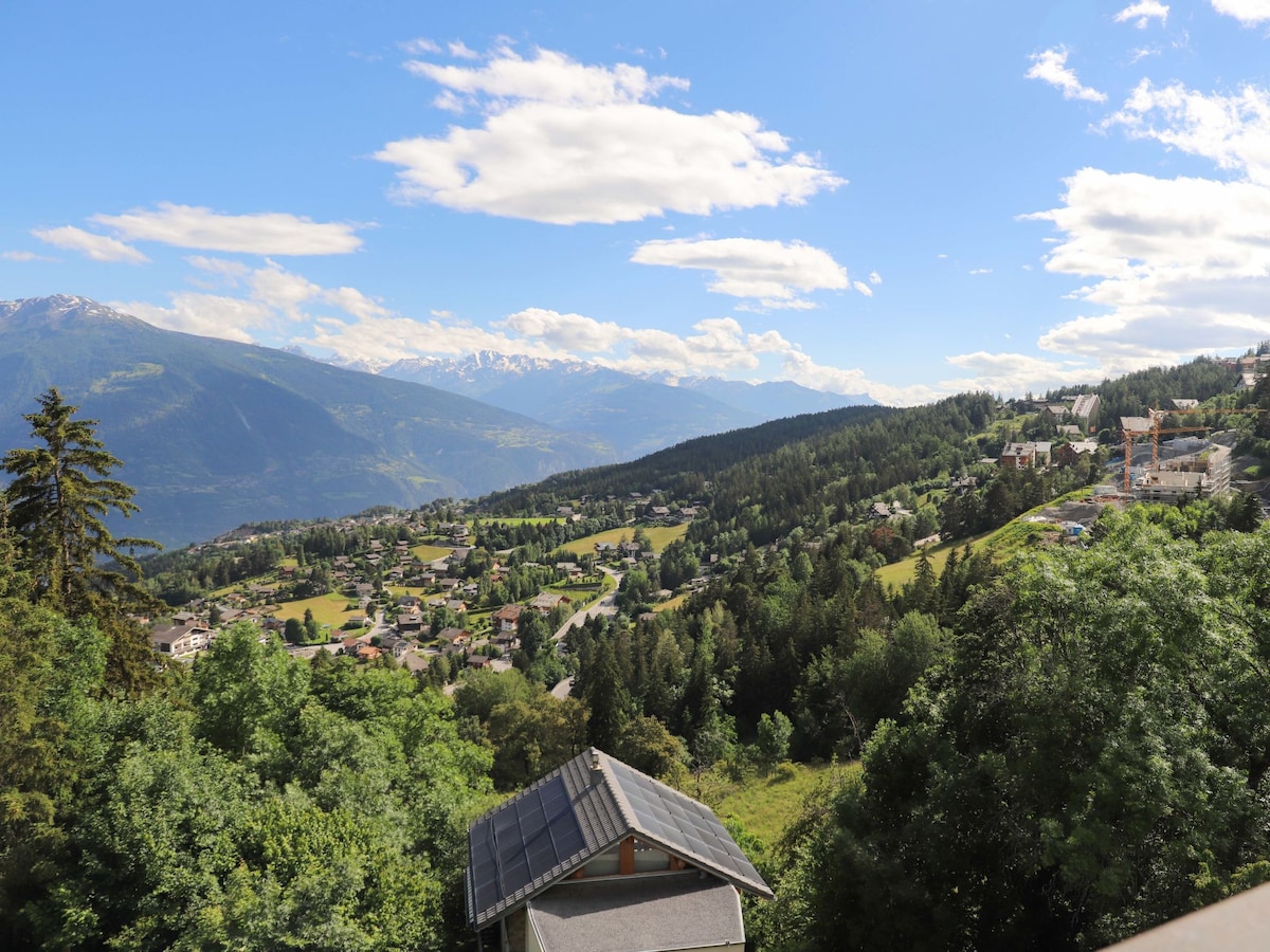 A panoramic view of the surrounding mountains and valley is presented, featuring lush green landscapes interspersed with residential areas. The sky is partly cloudy, with sunlight illuminating the scene, highlighting the natural beauty of the region.