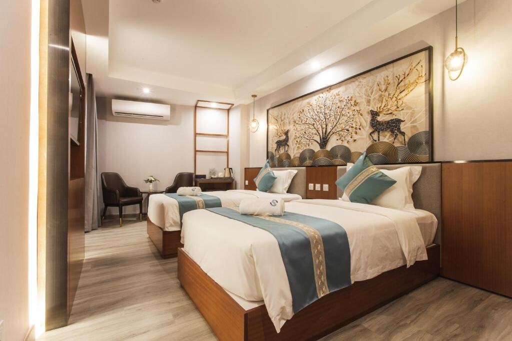 The room features two stylish twin beds, each with soft pillows and elegant blue accents. A decorative panel with artistic motifs is placed above the beds. Modern furnishings, including chairs and a work desk, complement the well-lit interior space.