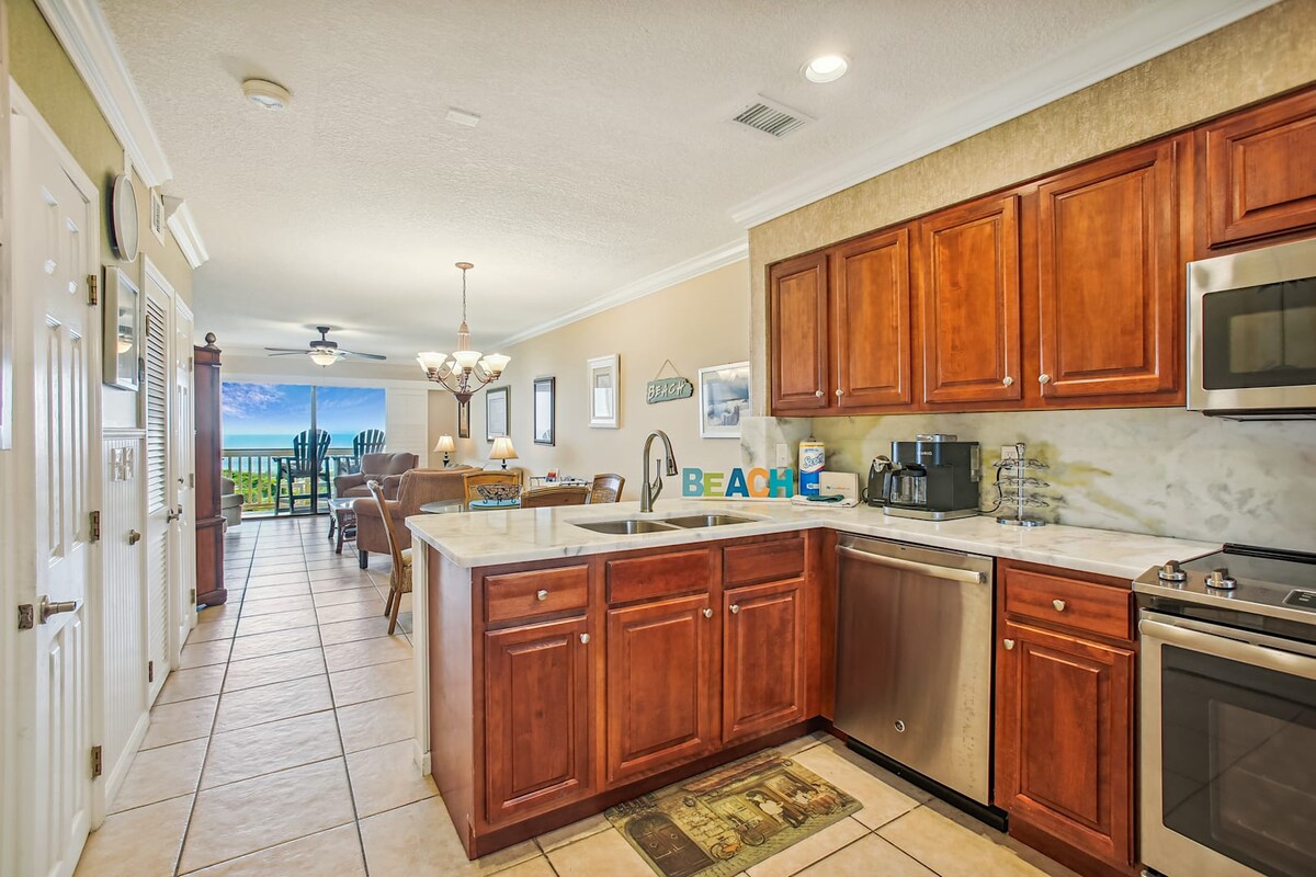 A well-equipped kitchen features wooden cabinetry and modern appliances, including a stainless steel refrigerator and dishwasher. The open floor plan leads to a dining area adorned with a chandelier, while large windows showcase views of the ocean beyond.