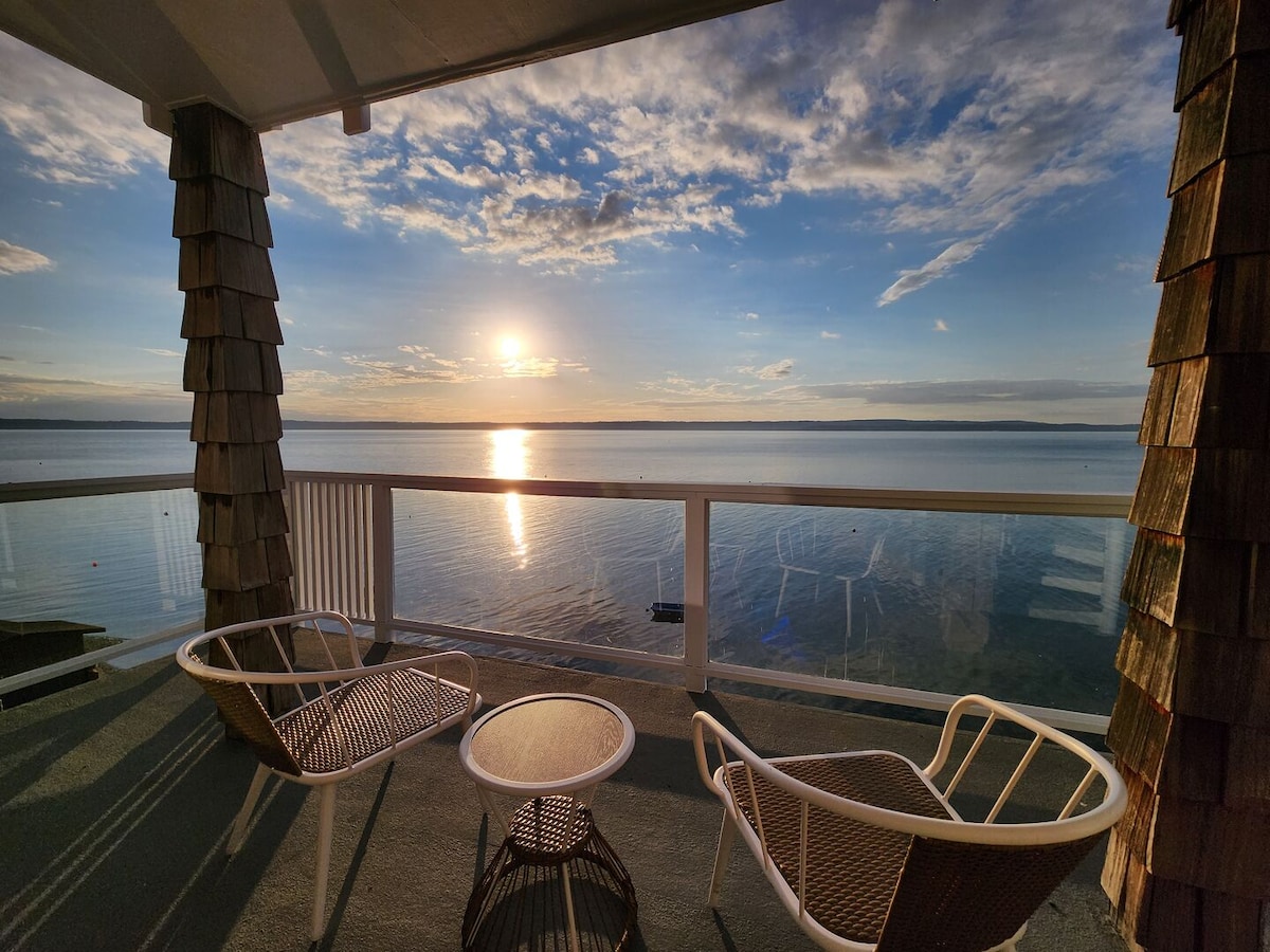 A covered outdoor deck features two chairs and a small table, with a breathtaking view of the calm water reflecting the sunset. The sun sets on the horizon, casting a warm glow over Port Susan Bay, framed by the peaceful shoreline.