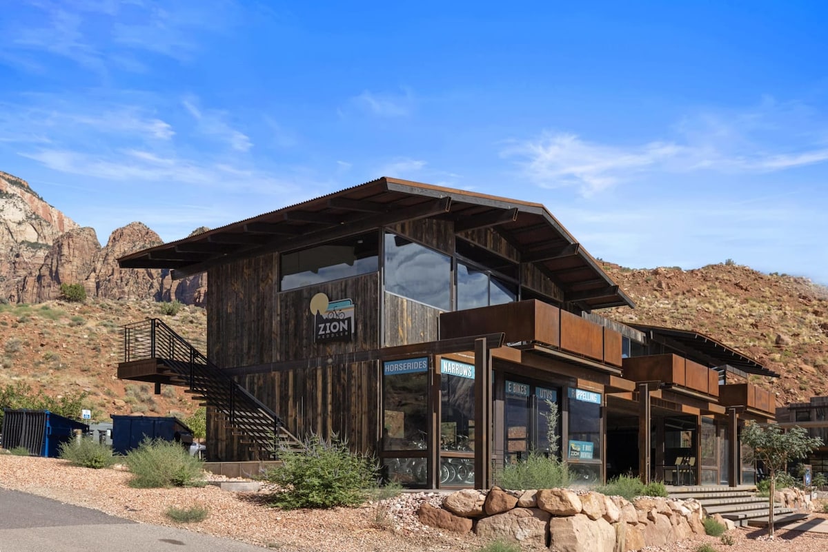 A modern building features a wooden exterior and expansive glass windows, offering views of the surrounding landscape. A staircase leads to the upper level, while desert vegetation is visible in the foreground. The location is enhanced by dramatic canyon cliffs in the background.