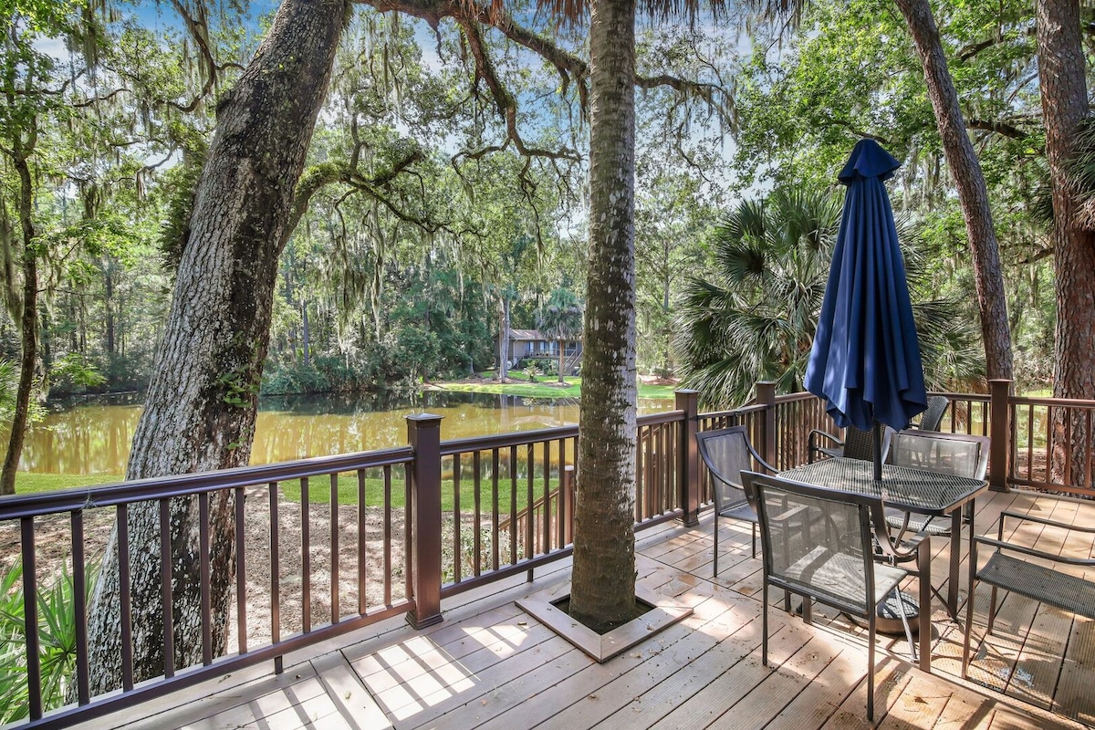 A spacious deck is shown with a dining set featuring four chairs and a table. A large umbrella provides shade, while lush trees frame the view overlooking a tranquil lagoon with natural surroundings.