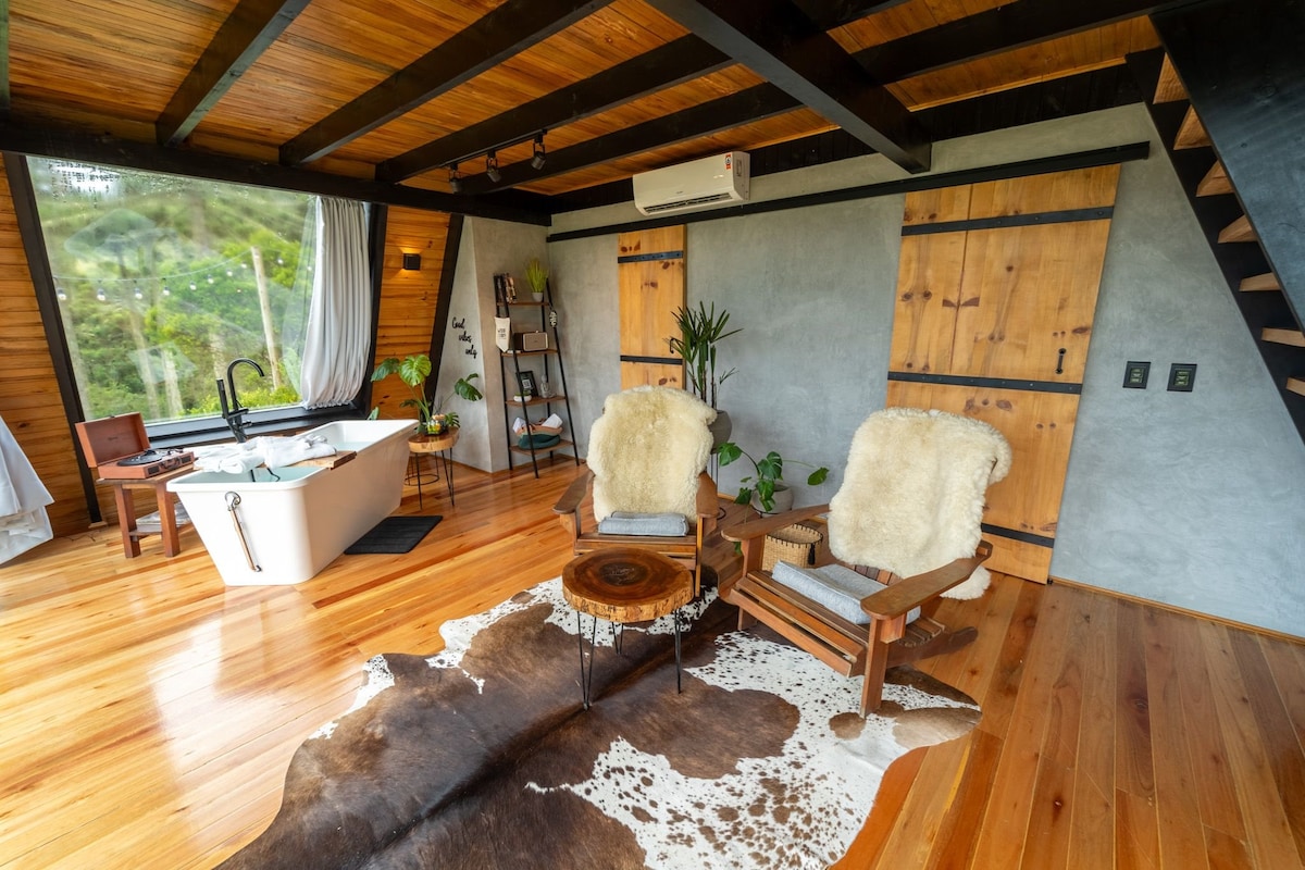 A welcoming interior features two wooden armchairs, each adorned with a soft throw, positioned beside a round wooden table. An inviting bathtub is visible nearby, complemented by natural light streaming through large windows that frame views of the mountains beyond.