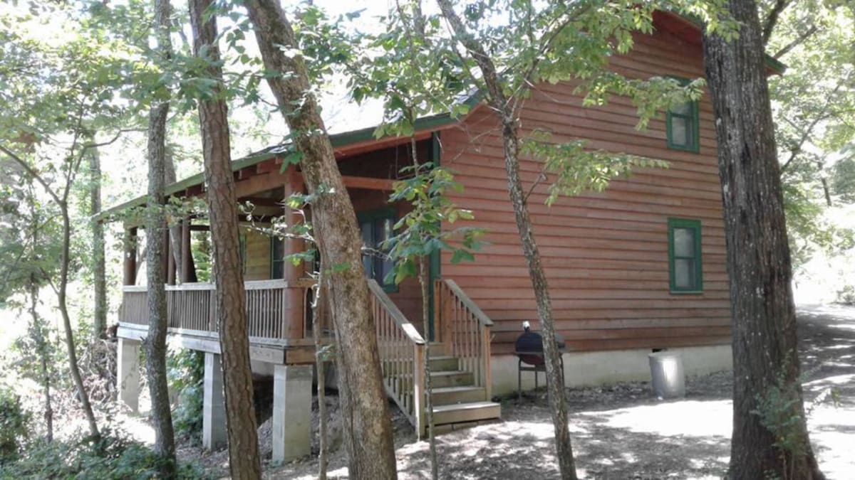 The cabin's exterior features rustic siding and knotty pine accents, surrounded by lush trees. A covered porch extends from the front, supported by log columns. Stairs lead up to the porch, which includes seating and a grill, offering a serene woodland atmosphere.