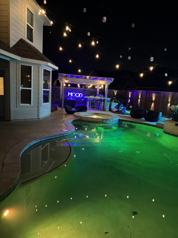 The outdoor oasis features a private pool surrounded by soft green lighting and illuminated by string lights overhead. A cozy seating area is visible, with a bar area in the background, all set against a serene night sky.