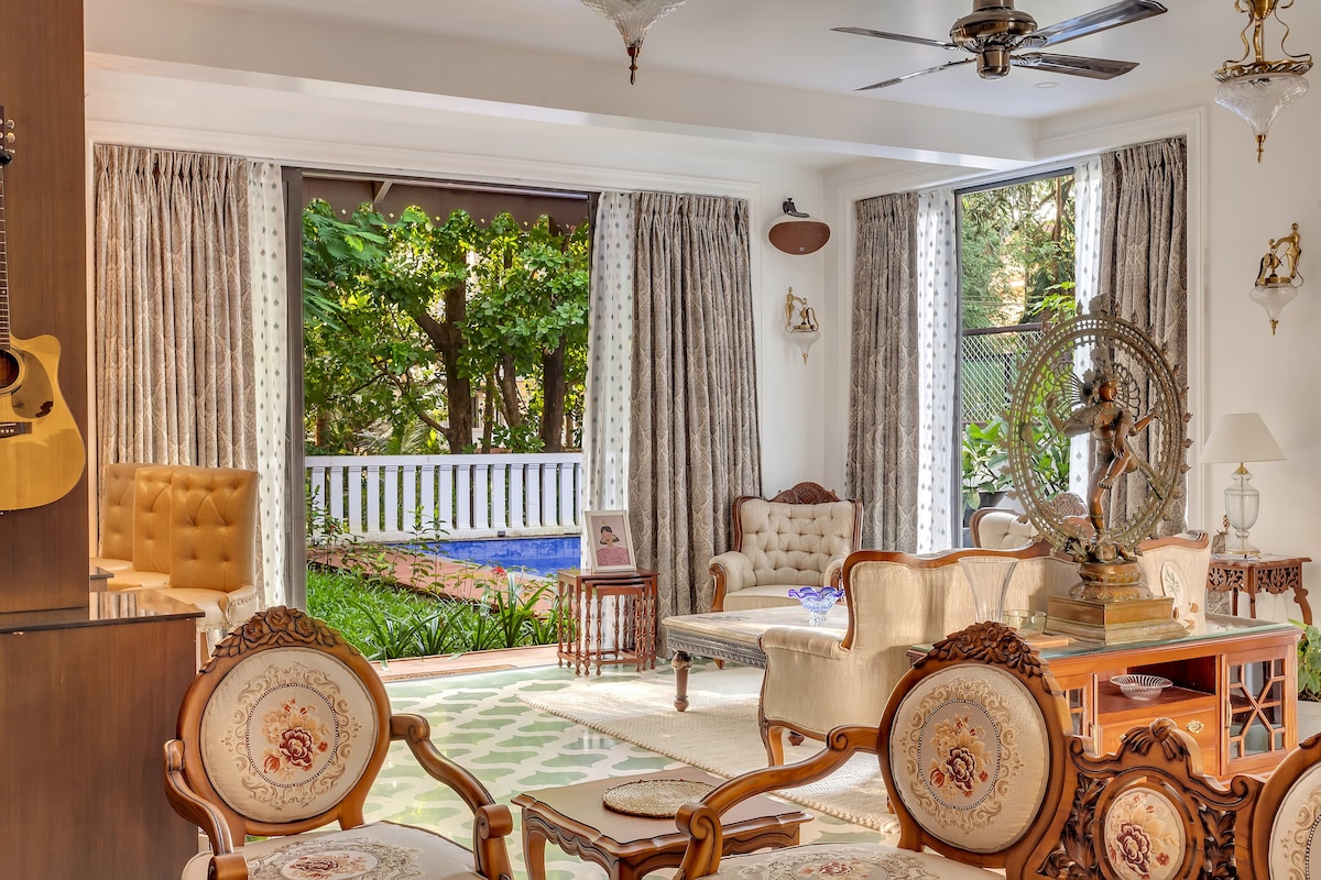 A spacious living room is showcased with elegant furniture, including a tufted chair and ornate sofa. Large windows frame views of lush greenery outside. Decorative elements, such as a guitar and a traditional statue, are present, enhancing the room's inviting ambiance.
