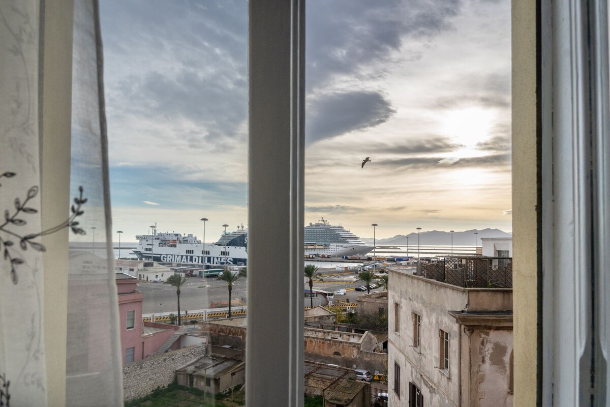 A window view showcases the harbor of Cagliari, featuring large cruise ships docked nearby. Soft curtains frame the scene, allowing natural light to illuminate the room. The sky is painted with clouds and hues of blue and gray, signaling a serene atmosphere.