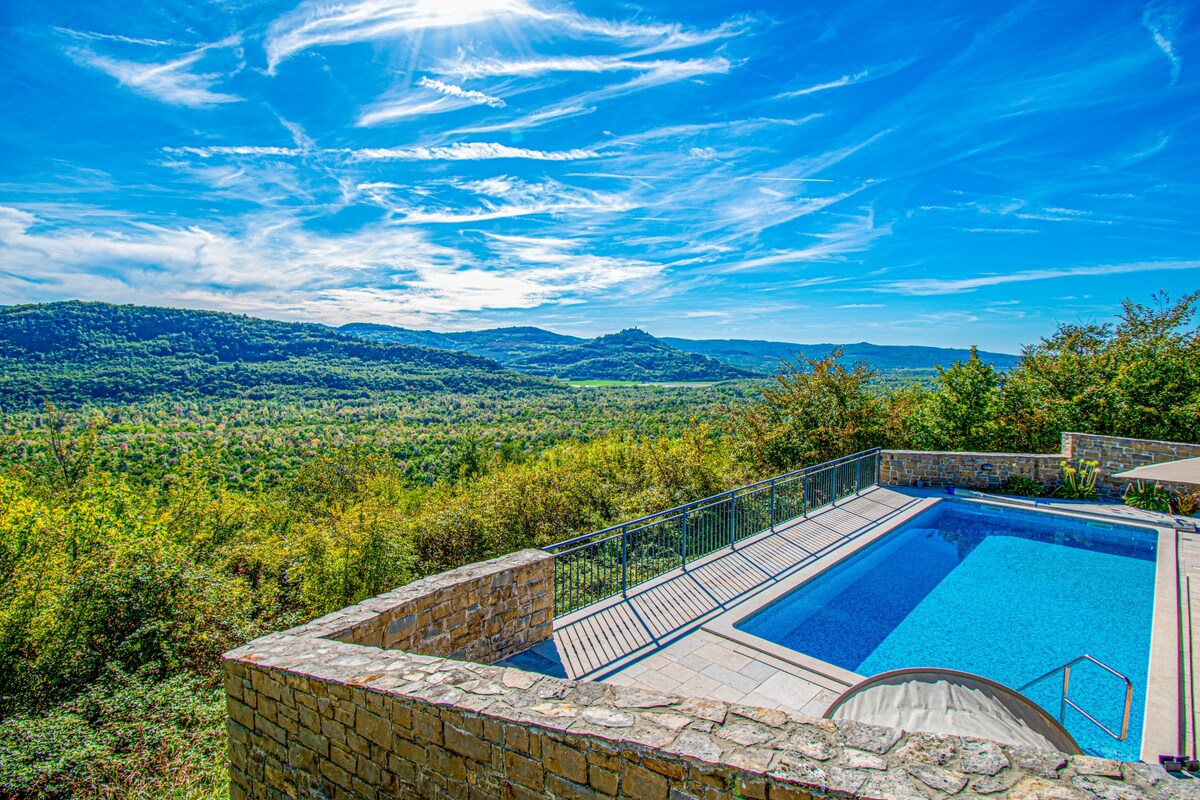 An outdoor pool is set within a stone patio, surrounded by lush greenery and rolling hills. Sunlight reflects off the water, and a clear blue sky is visible, enhancing the serene landscape. The area features a railing for safety and views of the picturesque surroundings.