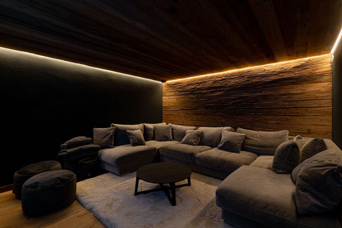 A cozy home cinema space features a spacious sectional sofa arranged around a circular coffee table. Warm light strips illuminate the wooden ceiling and accentuate the textured wooden wall. Soft poufs are positioned nearby on a plush area rug, creating an inviting setting for relaxation.