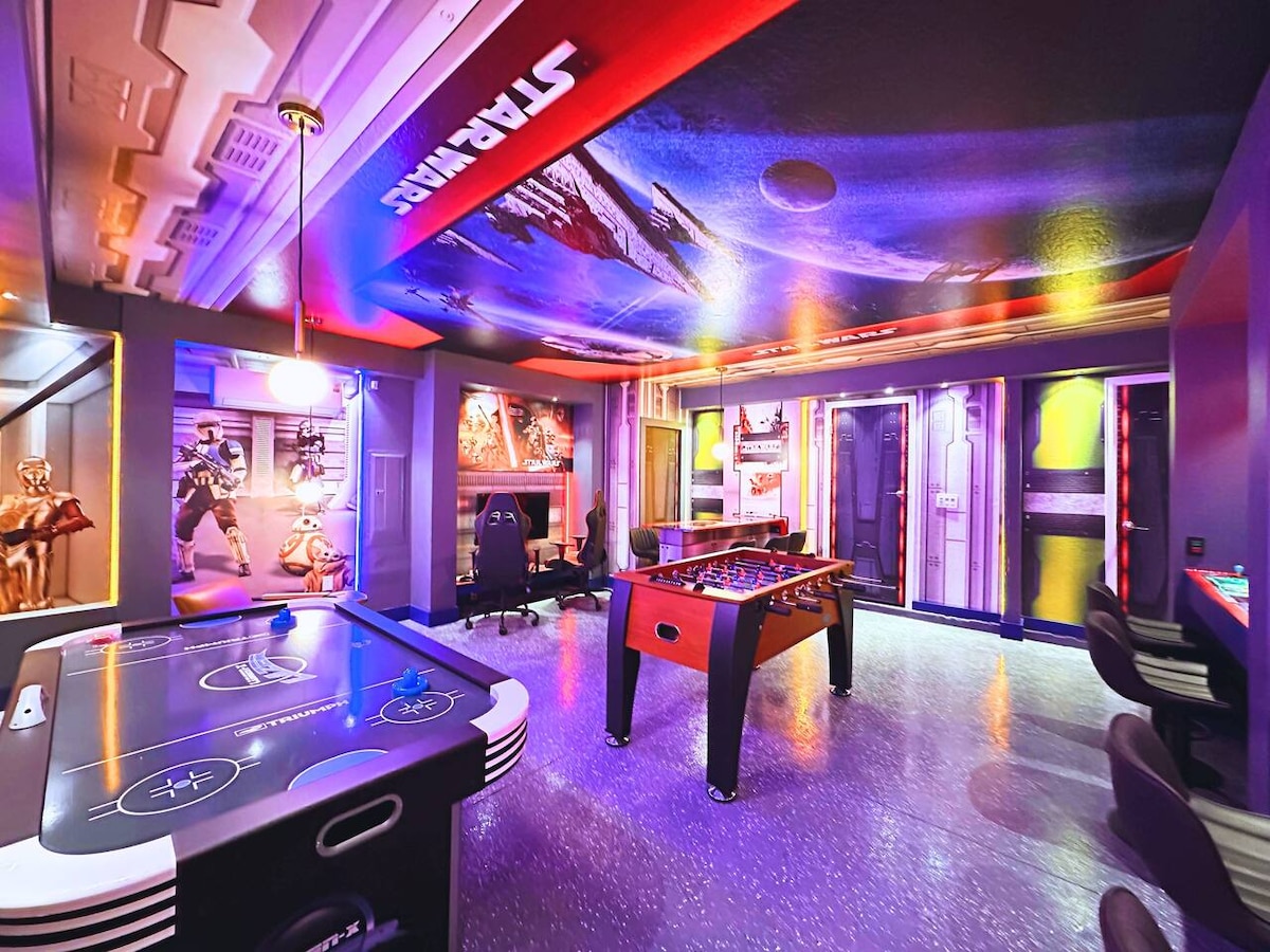 The game room features vibrant, themed decor with a striking Star Wars mural on the ceiling. An air hockey table and foosball table provide entertainment, while chairs and a gaming station are included for comfort and relaxation.