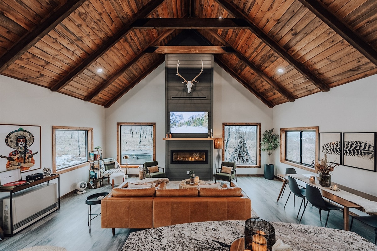 A spacious living room features large windows allowing natural light and partial mountain views. Cozy furniture surrounds a gas fireplace, complemented by a mounted deer head above. Hardwood ceilings enhance the rustic charm, while a dining table adds functionality to the area.