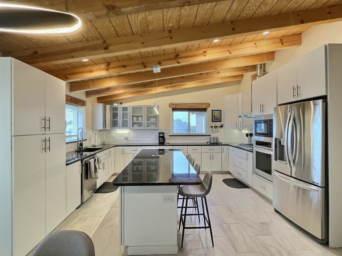 A modern kitchen features sleek cabinetry and high-end appliances. A spacious island with seating is positioned at the center, under wooden beams that add character. Ample natural light enters through windows, illuminating the open layout that seamlessly connects to the dining area.
